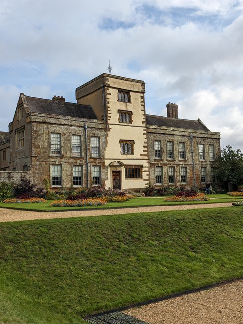 A large historic mansion with a symmetrical facade, constructed from stone and brick, featuring multiple rectangular windows with white frames, a central entrance with a small canopy, and a tower-like