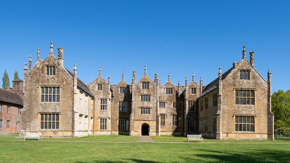 A large historic manor house with a symmetrical façade made of light-colored stone, featuring multiple windows with small panes, decorative stonework, and a central arched entrance. The building has