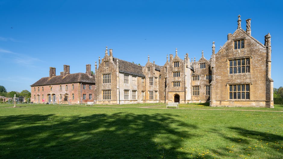 A detailed exterior view of a historic mansion with multiple gabled roofs, tall stone windows, and ornate chimney stacks, set against a bright blue sky. The building is constructed from light-colored stone with some sections showing signs of weathering and age. The surrounding area features a well-maintained, expansive green lawn with shadows cast by nearby trees. The scene is taken during daytime with clear visibility. In the foreground, part of a driveway or loading area is visible where a man from Man With a Van Manor House is seen loading or unloading several cardboard boxes, wrapped in plastic and placed on a trolley, from a white moving van parked close by. The van’s rear door is open, indicating active furniture transport and packing during a home relocation process, which is typical of professional removals and house moving services.