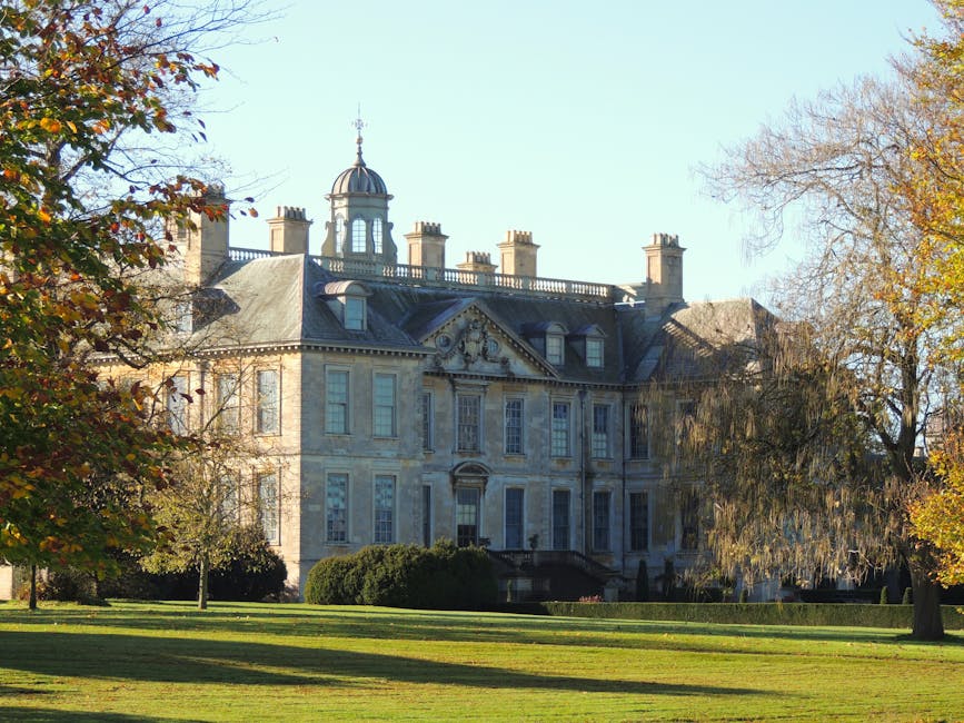 A large, historic mansion with a grey stone facade and multiple chimneys, situated behind a well-maintained lawn. The building features tall windows with white frames, a central entrance with steps leading up to it, and a decorative triangular pediment above the front door. The roof has a small domed tower with a weather vane on top. Surrounding the property are trees with autumn foliage, some with orange and yellow leaves and others mostly bare. In the foreground, a move involves a person loading cardboard boxes, wrapped in plastic and packing materials, into the back of a white moving van parked at the property’s entrance. The scene takes place during daytime with clear skies, and the environment suggests a home relocation or furniture transport process managed by a moving service, such as Man With a Van Manor House.