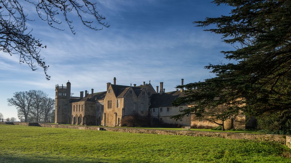 A historic stone castle or manor house situated on a grassy landscape with a clear blue sky overhead. The building features multiple turrets, chimneys, and gabled roofs, with large arched windows and stone walls. Surrounding the structure are mature leafless trees on the left and evergreen trees on the right, partially framing the scene. The foreground shows a well-maintained lawn and a stone pathway or boundary wall, indicating the property’s extent. The scene is illuminated by natural daylight, highlighting the architectural details and casting mild shadows, suggestive of a peaceful, rural setting suitable for home relocation or moving services by Man With a Van Manor House.