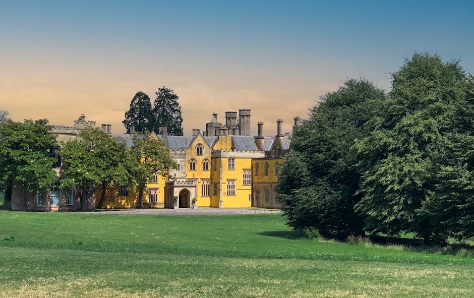 A large, historic yellow mansion with multiple turrets, chimneys, and decorative windows situated behind a green lawn and several mature trees. The building appears to be part of a property undergoing a home relocation or furniture transport process, typical of professional removals services. The mansion is visible from a distance, with the surrounding environment including open grass and leafy trees framing the building. The lighting suggests late afternoon or early evening, with a clear sky above. This scene likely depicts the exterior setting before the moving team begins packing, loading, or transporting household items, demonstrating the property’s spacious surroundings and the scale of the building involved in a house removal operation handled by Man With a Van Manor House.