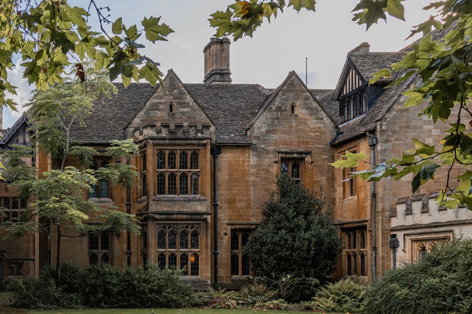 The image shows a large, historic-style stone house with multiple gabled roofs and tall, narrow windows featuring leaded glass panes, situated within a garden with green foliage and trees. The house's facade is constructed from warm-colored sandstone, giving it a classic and stately appearance. Surrounding the building are leafy branches and trees, some of which partially obscure the structure, suggesting an outdoor setting with natural lighting. In the foreground, there appears to be a paved driveway or pathway, possibly used for vehicle access and parking, which would be relevant during home relocation or furniture transport activities. The scene emphasizes the traditional architecture and greenery typical of residential properties in Manor House, appropriate for discussing moving and packing logistics, including loading and unloading furniture, boxes, and packing materials like cardboard and plastic wrap. Occasionally, Man With a Van Manor House assists with such house removals, ensuring compliance with local parking and moving permits as outlined by Hackney Council rules for Manor House.