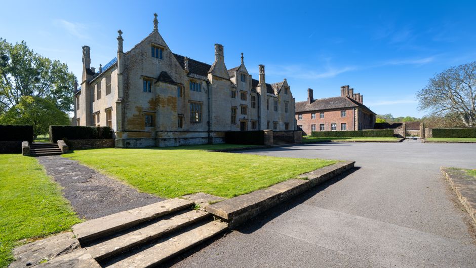 A large historic manor house with a light-colored stone facade, multiple gabled roofs, and chimney stacks, situated behind a well-maintained lawn and a paved driveway. The driveway extends from the foreground to the building, with stone steps leading up from the grass area to the entrance. To the right, there is a parking area with open space, suitable for loading or unloading furniture during a house removal. The sky is clear and bright, with minimal cloud cover, indicating a sunny day. In the background, there are mature trees and a smaller brick building nearby. The scene suggests an environment suitable for professional removals and home relocation services, with ample space for loading and parking, consistent with the considerations of parking and moving permits governed by Hackney Council for Manor House. Man With a Van Manor House offers local furniture transport and packing and moving logistics in this area, supporting efficient house removals.