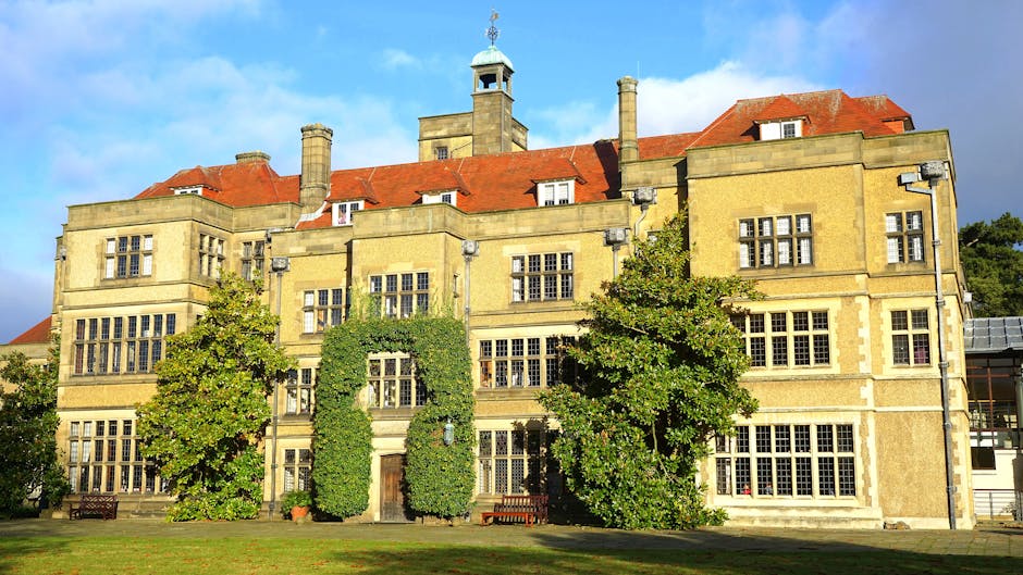 A large historic building with a beige stone exterior, multiple rectangular windows, and a red-tiled roof, situated behind a lawn with several mature trees. The facade features decorative stonework around the windows and central doorway, partially obscured by green ivy climbing up the walls. Visible in the foreground are the paved area and the grass, highlighting the building's exterior and outdoor surroundings. This scene illustrates the type of property that might require professional house removals or furniture transport services. Man With a Van Manor House, a relocation company, could facilitate packing and moving operations in this area, especially when managing home relocation logistics for such sizeable historic properties, including loading household furniture and boxes onto vans for transport from Green Lanes flats or other nearby locations.