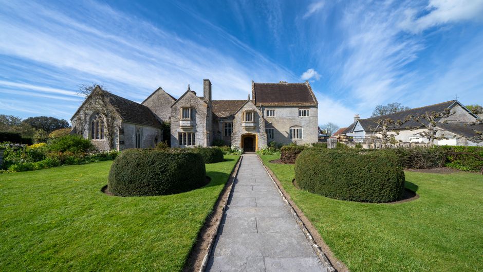 A stone driveway leading directly to the entrance of a large, historic manor house with a pitched roof and multiple gothic-style windows. The driveway is lined with neatly trimmed, rounded bushes on either side, set within a well-maintained lawn. The front door of the manor house is visible at the end of the pathway, framed by an archway. The house is situated in a rural or suburban setting, with additional buildings and trees visible in the background under a bright, blue sky with wispy clouds. This scene exemplifies a residential property undergoing or prepared for a home relocation or moving process, with a focus on clear access for furniture transport and logistics support from companies like Man With a Van Manor House.