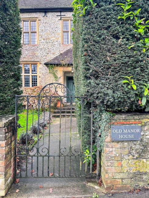 A black wrought iron gate with curved decorative elements, partially open and leading to the front entrance of an old stone manor house. The gate is set between low brick pillars and is surrounded by dense, well-maintained greenery, including trimmed hedges and climbing plants. The stone path behind the gate extends toward stone steps that ascend to a green front door with a small overhanging roof. To the right of the gate, a grey signage plaque reads 'OLD MANOR HOUSE,' mounted on a brick pillar. The building above features tall, multi-pane sash windows with wooden frames, and the exterior walls are constructed from weathered stone blocks. The scene is lit naturally, suggesting daytime, and depicts the entrance area of a traditional residential property, relevant for house removals or relocation logistics with a focus on the exterior and access point.