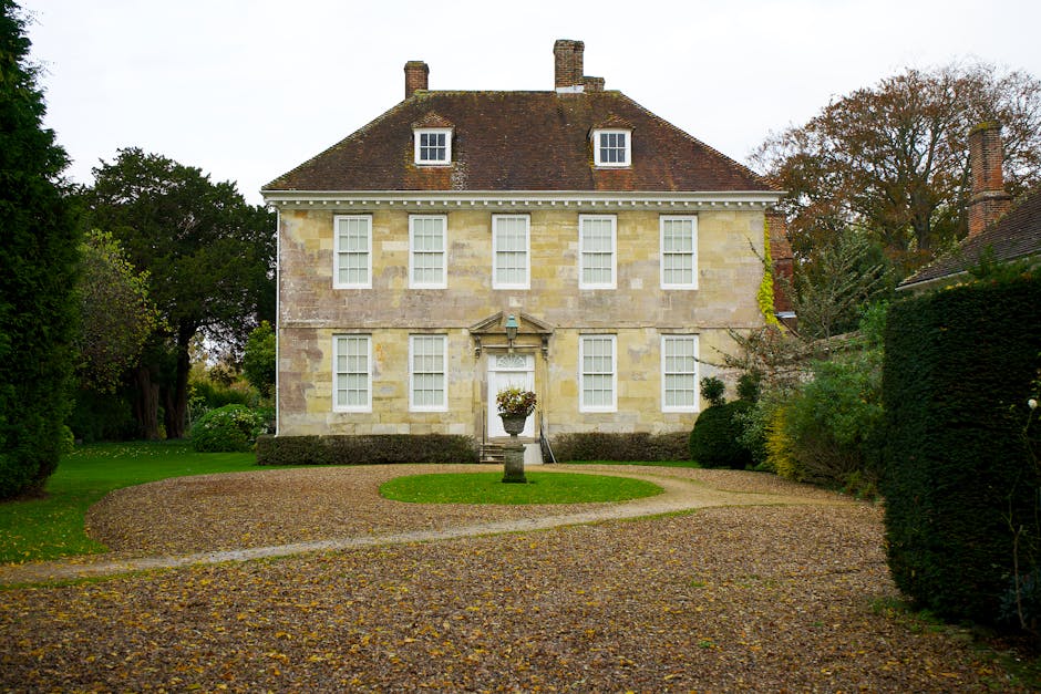 A stone driveway leading directly to the entrance of a large, historic manor house with a pitched roof and multiple gothic-style windows. The driveway is lined with neatly trimmed, rounded bushes on either side, set within a well-maintained lawn. The front door of the manor house is visible at the end of the pathway, framed by an archway. The house is situated in a rural or suburban setting, with additional buildings and trees visible in the background under a bright, blue sky with wispy clouds. This scene exemplifies a residential property undergoing or prepared for a home relocation or moving process, with a focus on clear access for furniture transport and logistics support from companies like Man With a Van Manor House.