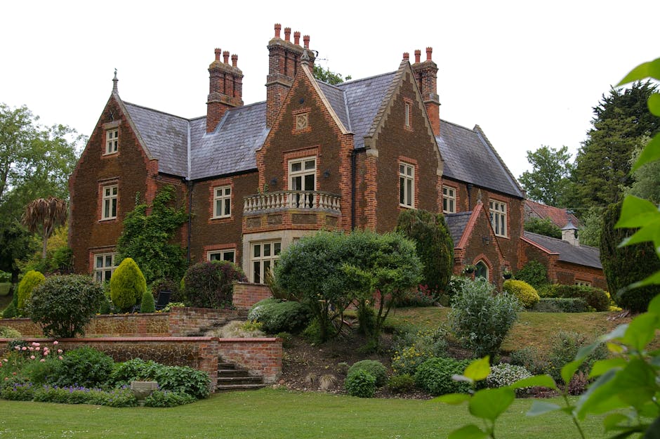 A large, historic brick house with multiple gabled roofs, tall chimneys, and decorative stonework, situated in a lush, landscaped garden with various shrubs, trees, and flowering plants. The house features white-framed windows and a small balcony on the upper floor. In the foreground, there is a paved pathway and a brick retaining wall. Outside the house, a man from Man With a Van Manor House is seen loading or unloading boxes and furniture wrapped in plastic and blankets from a large van parked on the pavement, indicating a home relocation or furniture transport process as part of professional removals services.