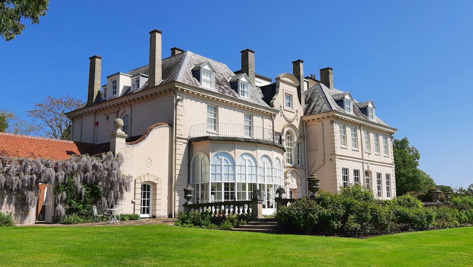 A large, elegant multi-storey house with a cream-colored exterior and a slate roof, situated against a clear blue sky. The building features multiple chimneys, bay windows with white frames, and intricate architectural details. The front garden is well-maintained with a lush green lawn, small bushes, and a flowering plant with cascading purple blossoms on the left side. In the foreground, outside the property near the pavement, a man from Man With a Van Manor House is involved in a home relocation process, loading cardboard boxes, furniture wrapped in plastic and moving blankets, and other packing materials into a van. The van parked adjacent to the driveway is partially visible, along with moving equipment such as trolleys and straps, indicating an organized furniture transport and packing operation for house removals and professional moving services near Clissold Park.