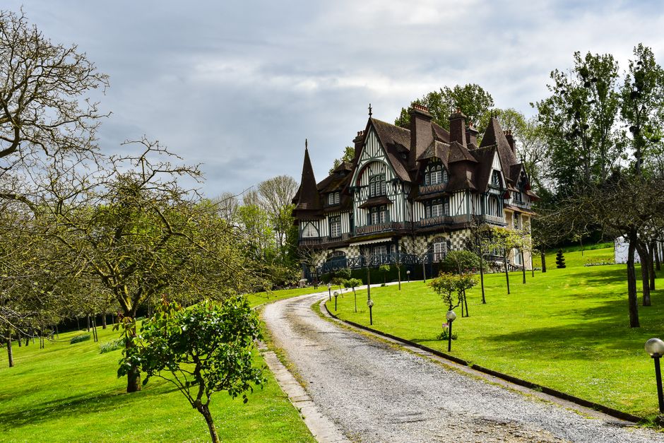 A long, curved gravel driveway leading up to a large, Victorian-style manor house with multiple gabled roofs, ornate dark wood and white exterior features, and tall chimneys. The driveway is bordered by well-maintained green lawns, several small trees, and evenly spaced lampposts. The scene is set outdoors during daytime with a partly cloudy sky, and surrounding the property are mature trees with some leafless branches and others with fresh green foliage. The image depicts a peaceful residential setting, suitable for home relocation or moving services, with the grand house situated at the end of the driveway accessible for furniture transport and packing during house removals, as handled by Man With a Van Manor House.