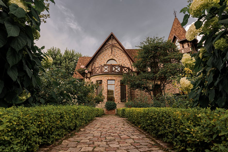 A long, curved gravel driveway leading up to a large, Victorian-style manor house with multiple gabled roofs, ornate dark wood and white exterior features, and tall chimneys. The driveway is bordered by well-maintained green lawns, several small trees, and evenly spaced lampposts. The scene is set outdoors during daytime with a partly cloudy sky, and surrounding the property are mature trees with some leafless branches and others with fresh green foliage. The image depicts a peaceful residential setting, suitable for home relocation or moving services, with the grand house situated at the end of the driveway accessible for furniture transport and packing during house removals, as handled by Man With a Van Manor House.