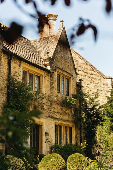Exterior view of a historic stone house with a steeply pitched roof and multiple chimneys, partially framed by blurred tree branches in the foreground. The house features yellow-painted window frames and a small outdoor lantern near the entrance. Surrounding the building are well-maintained bushes and greenery, suggesting a residential property. This image captures the traditional architecture often seen in home relocation or house removals contexts, with natural lighting highlighting the textured stonework. The setting is calm and residential, appropriate for content related to house removals, packing, and logistical planning for moving services like those offered by Man With a Van Manor House.