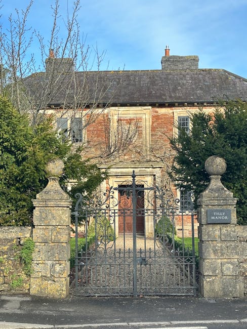 Photograph of Tilly Manor, an old brick house with a pitched roof, partially obscured by leafless trees and green shrubs. The building features a central stone doorway with an arched wooden door, framed by weathered stone detailing. In the foreground, a black wrought iron gate with ornate scrollwork is closed and attached to two stone pillars, each topped with a rounded stone finial, indicating the property boundary. The scene is set outdoors on a clear day with bright blue sky and sunlight illuminating the house and surrounding greenery. This image relates to house removals and moving logistics, as Man With a Van Manor House offers professional relocation services, including clearing and disposal of bulky items, within the context of home relocations and furniture transport.